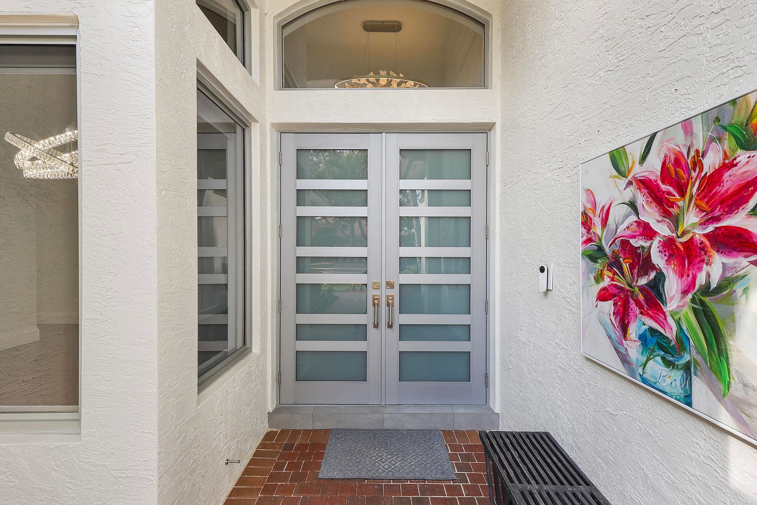This is a well-lit entryway featuring double doors with frosted glass panels and a transom window above. The exterior is painted in a light color, contrasting with the brick flooring and a modern bench. A piece of floral art adds a pop of color to the scene, creating a welcoming and stylish entrance.