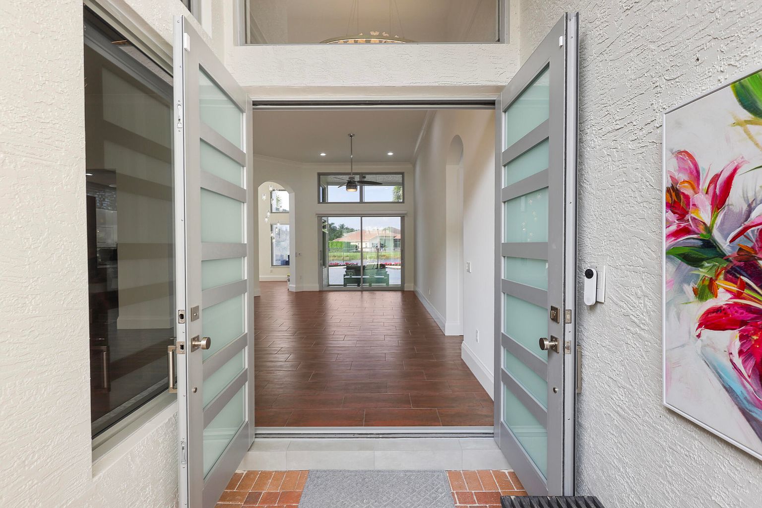 The image showcases an inviting entryway with double doors open to reveal a glimpse of the interior. The doors feature a modern design with frosted glass panels and sleek frames. The flooring transitions from an exterior brick and stone to an interior wood, creating a seamless flow. A painting is visible on the right wall, adding a touch of elegance.