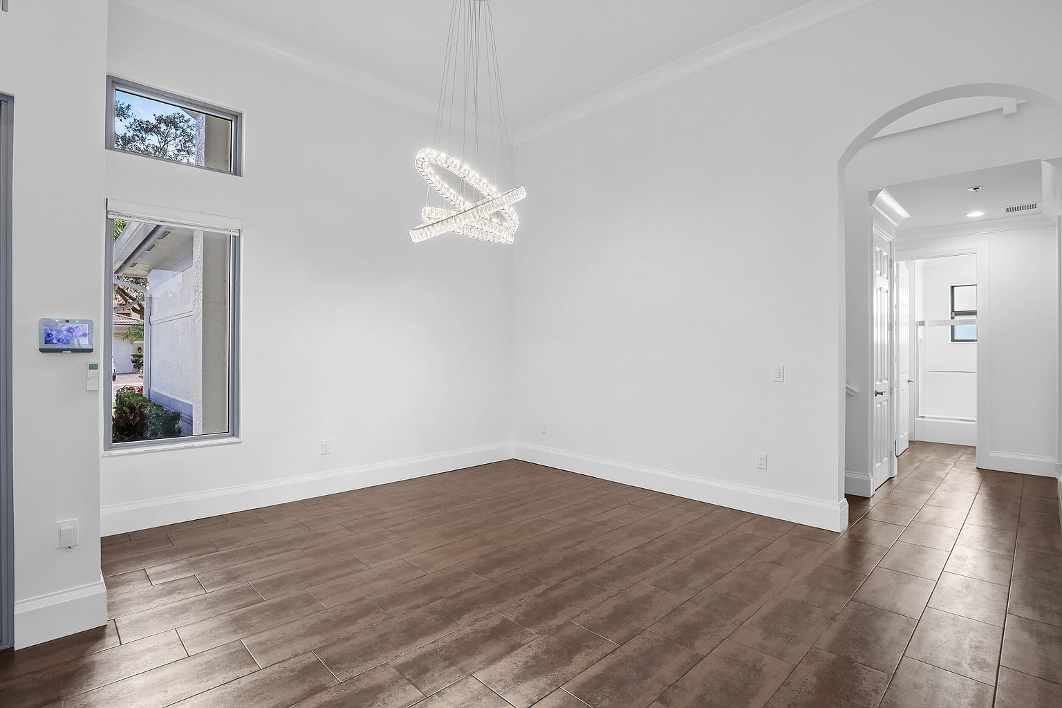 This is an interior shot of a dining room featuring dark wood-look tile flooring and white walls. A modern, abstract chandelier hangs from the ceiling, providing a focal point. An arched doorway leads to another room, adding depth to the space, and windows provide natural light.