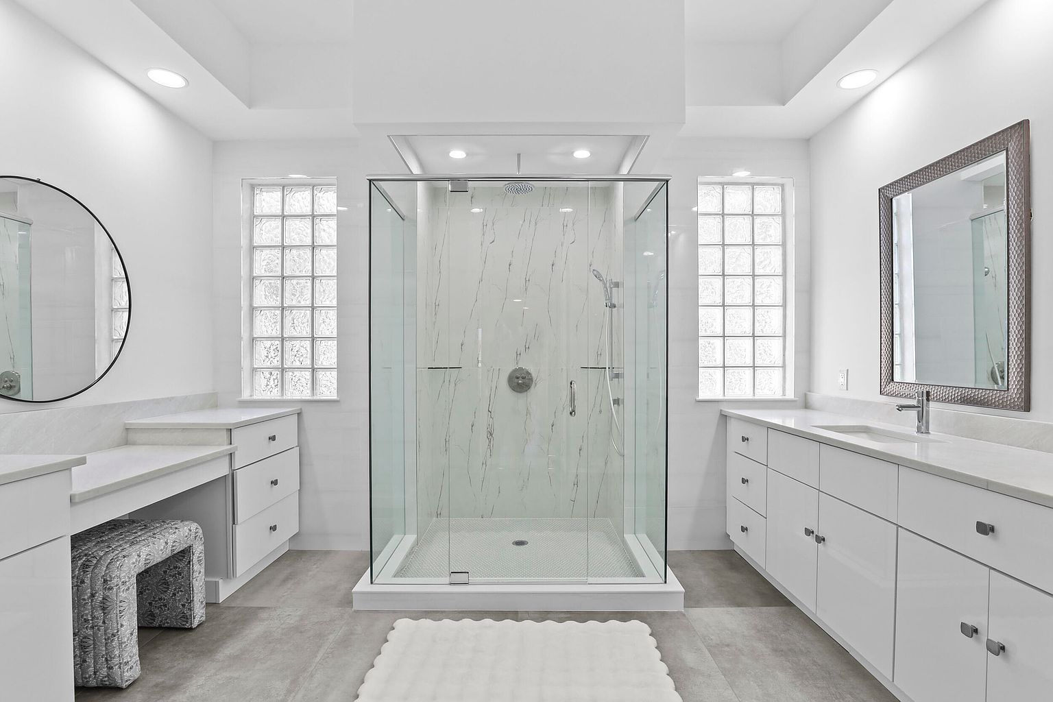 This is a bright and modern primary bathroom featuring a glass-enclosed shower with white marble-patterned walls and a gray tiled floor. The bathroom includes dual vanities with white cabinets and countertops, complemented by a round mirror on one side and a rectangular mirror on the other. Natural light streams in through glass block windows, enhancing the clean and spacious feel of the room.