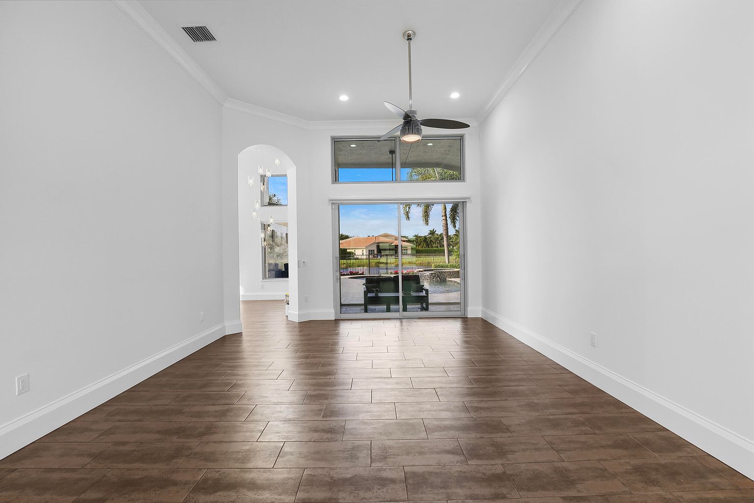 This is an interior shot of a dining room featuring dark wood-look tile flooring and white walls. A large sliding glass door provides a view of the outdoor pool and landscaping, while a ceiling fan hangs above. The room is bright and spacious, creating an inviting atmosphere.