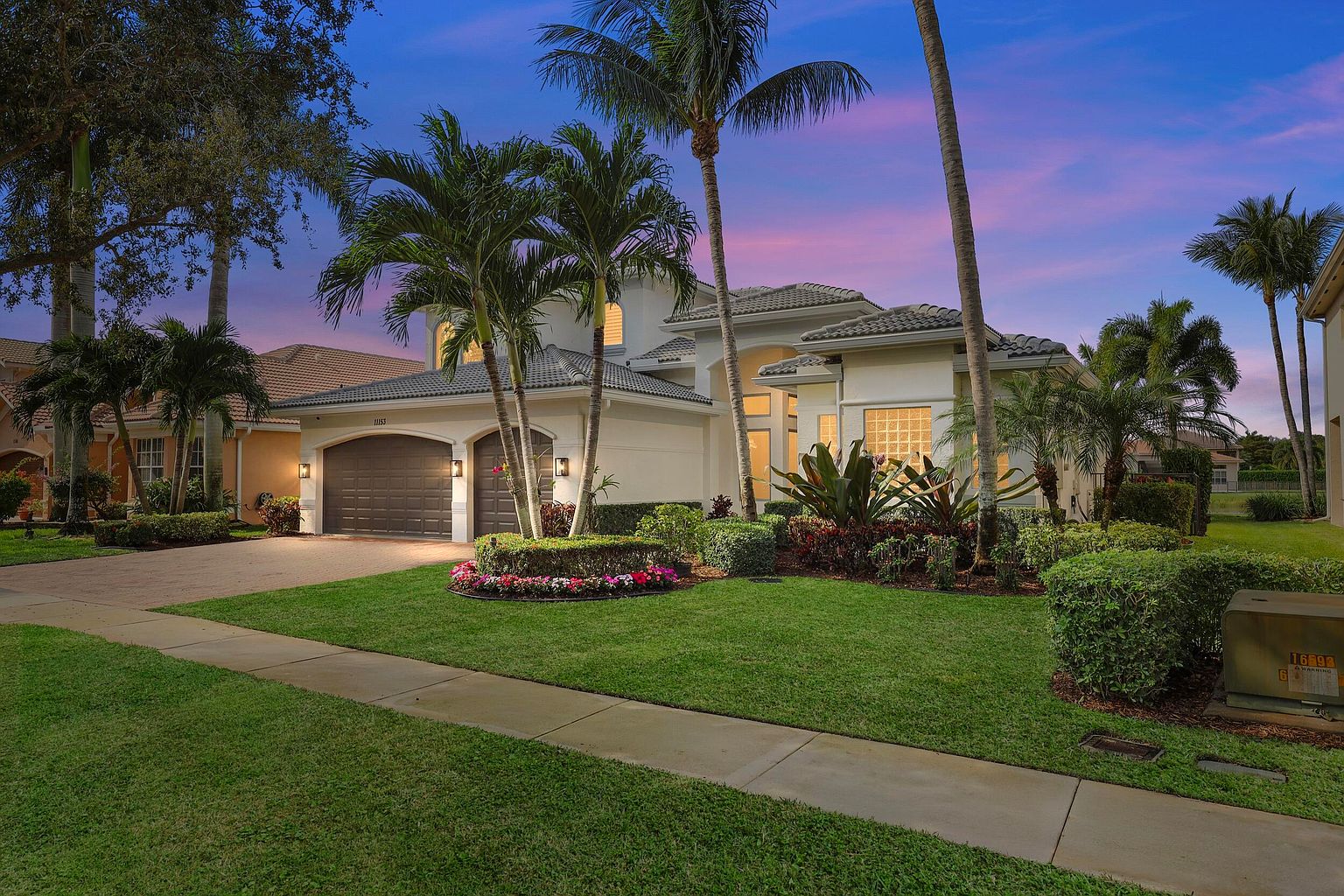 This is a front exterior view of a well-maintained, single-family home. The house features a two-car garage, a manicured lawn, and mature palm trees in the front yard. The architecture is modern with a neutral color palette, and the overall impression is one of luxury and curb appeal.