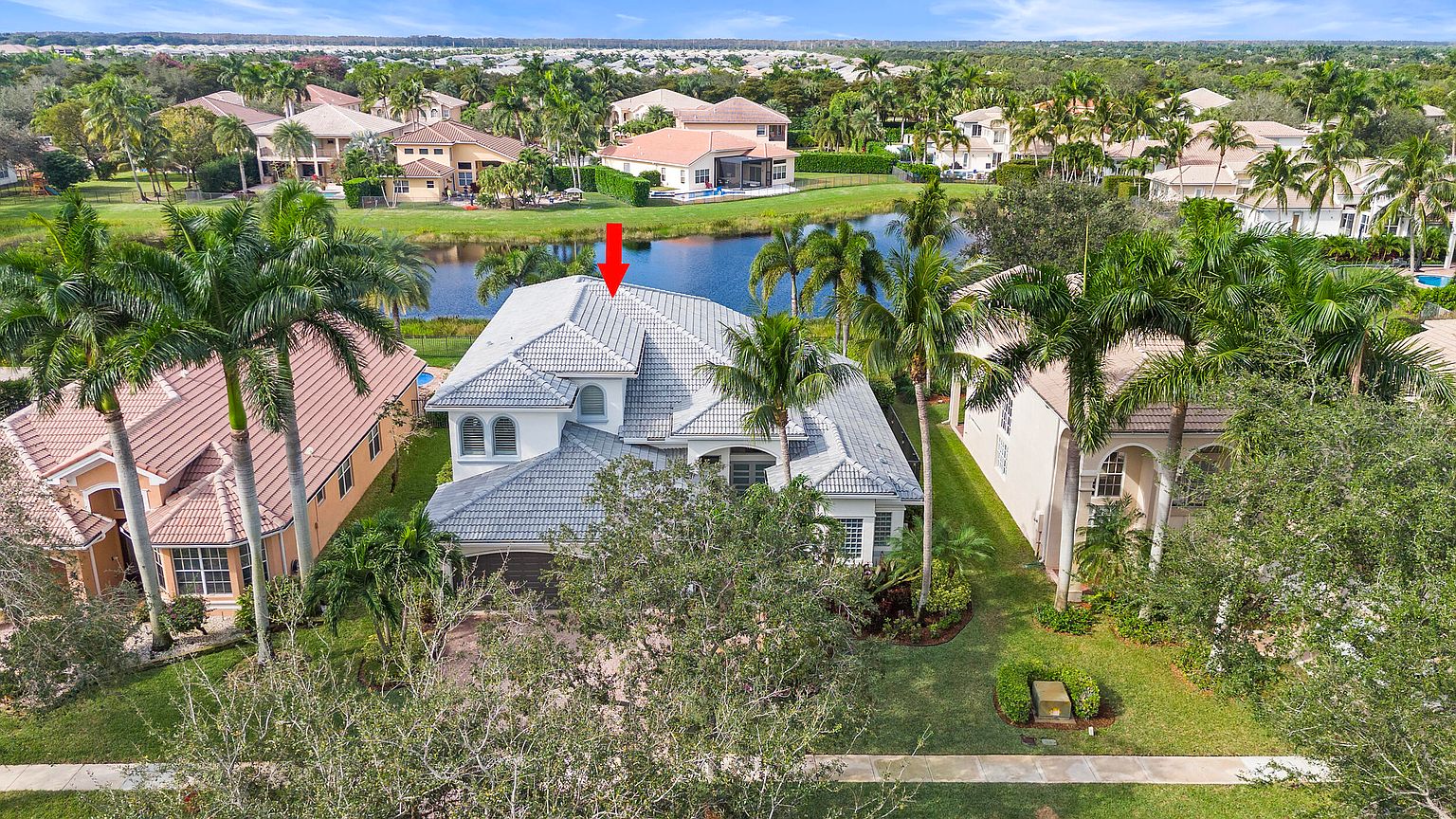 This aerial shot showcases a luxurious home with a gray tiled roof, surrounded by lush landscaping and a serene lake. The property features well-manicured lawns, mature palm trees, and a glimpse of neighboring houses, creating a sense of community within a tropical setting. The overall impression is one of upscale living and tranquility.