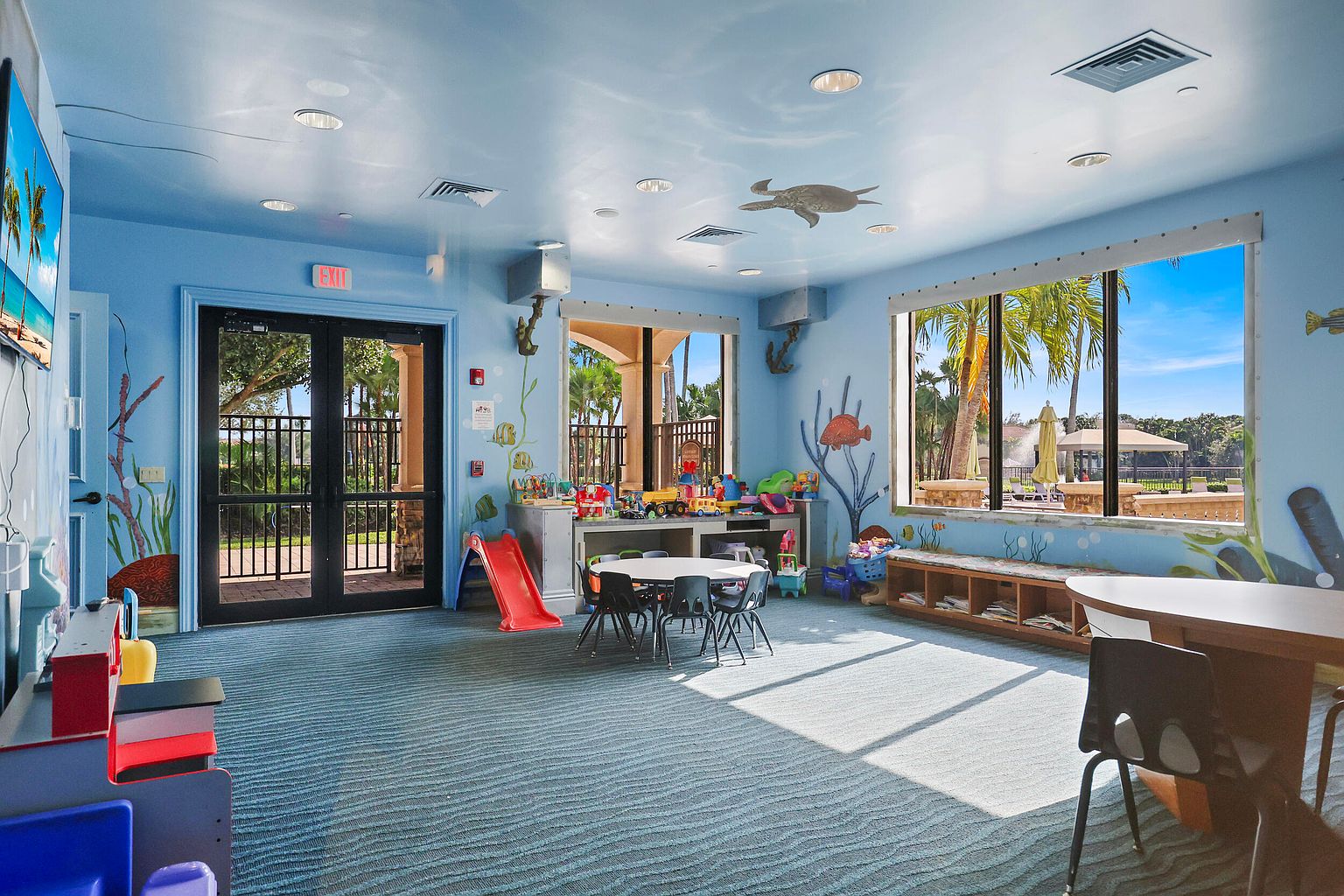 This is an interior shot of a children's playroom in a community amenity space. The room is brightly colored with blue walls and a patterned blue carpet, featuring murals of sea life. There are various toys, tables, and seating arrangements, with natural light streaming in from large windows overlooking an outdoor area.
