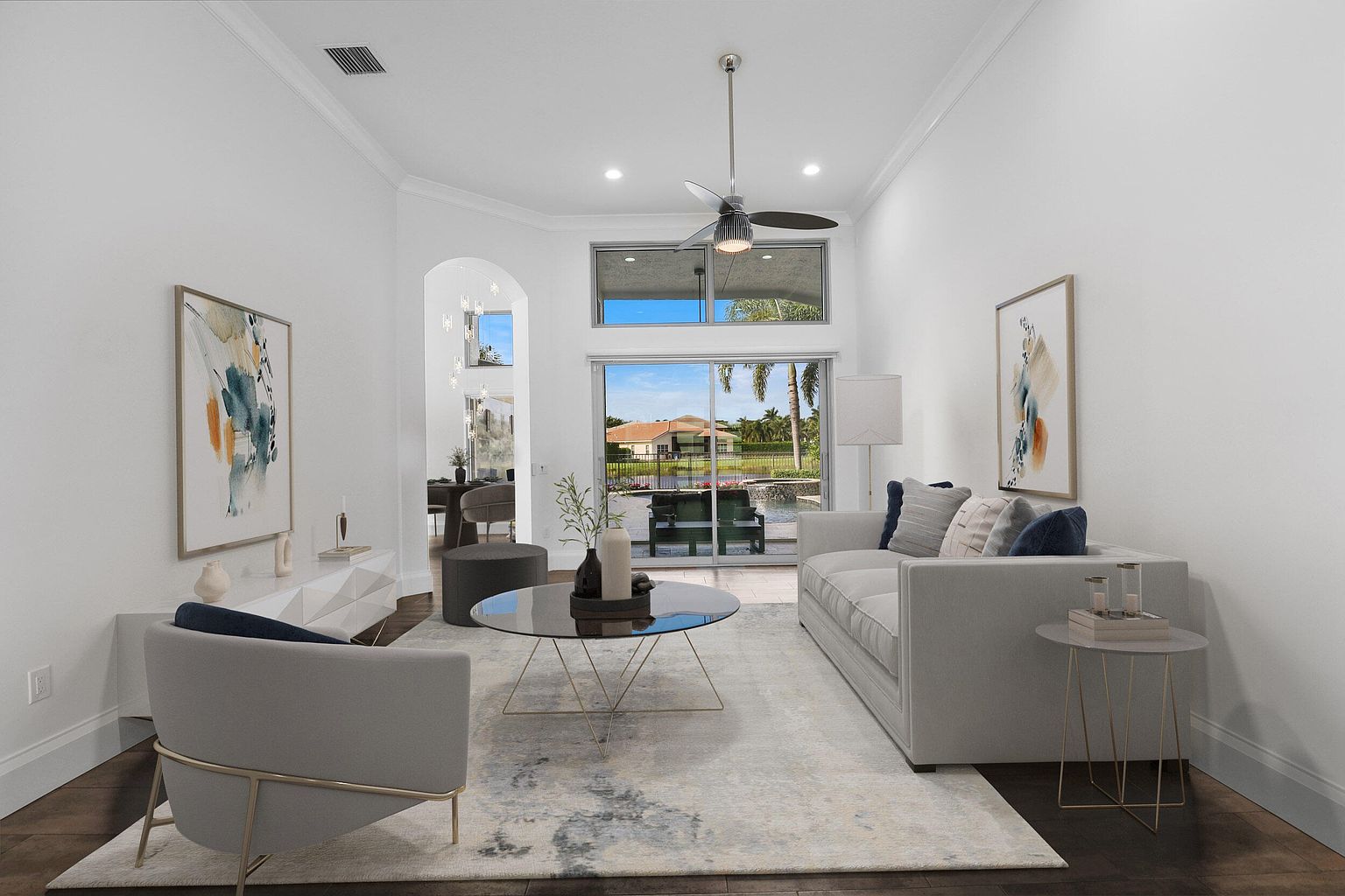 This is an interior shot of a living room, showcasing a bright and airy space with modern furnishings. The room features a light-colored sofa, a glass-topped coffee table, and artwork on the walls. Large windows provide a view of the outdoors, enhancing the room's appeal and creating a serene atmosphere.