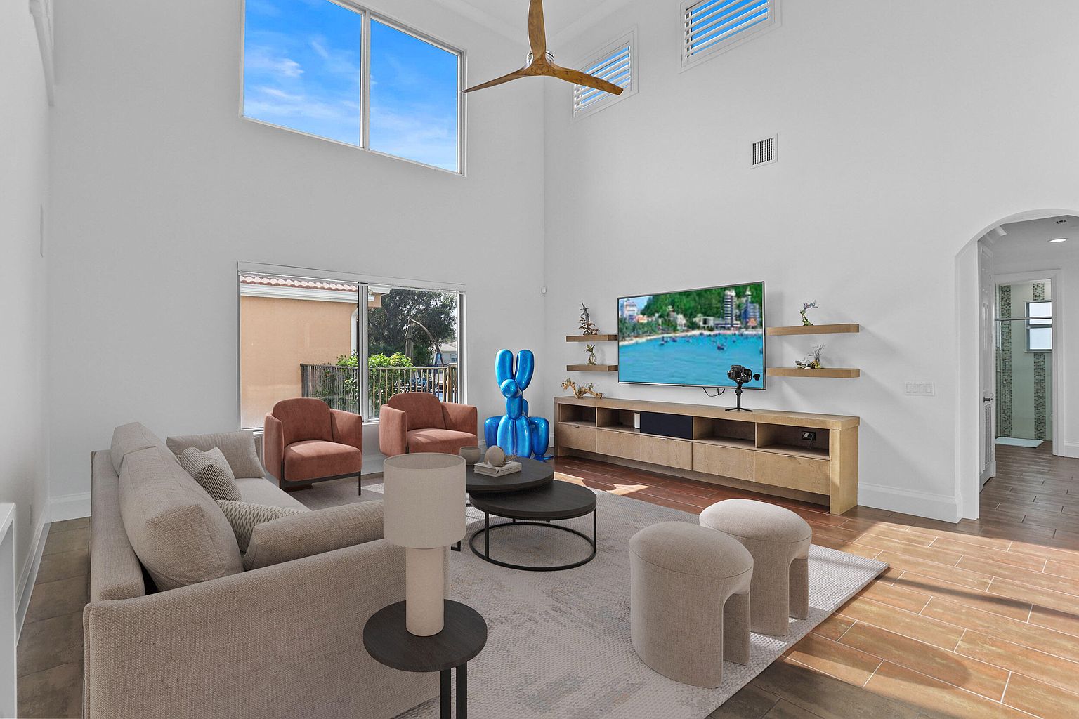 This is a bright and airy living room with high ceilings and large windows that provide ample natural light. The room features a neutral color palette with a comfortable sofa, two armchairs, and modern decor. A large television is mounted on the wall above a wooden media console, and the hardwood flooring adds warmth to the space.