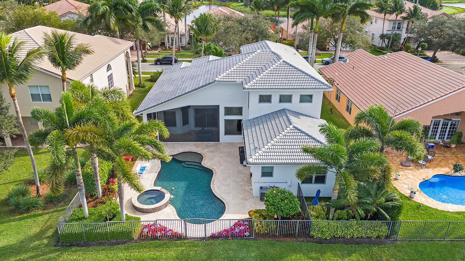 This aerial view showcases a luxurious backyard featuring a freeform pool and a circular spa, surrounded by a paved patio. The property is enclosed by a decorative metal fence and lush landscaping, including palm trees and flowering plants. The house has a modern architectural style with a gray tiled roof and a screened-in porch.