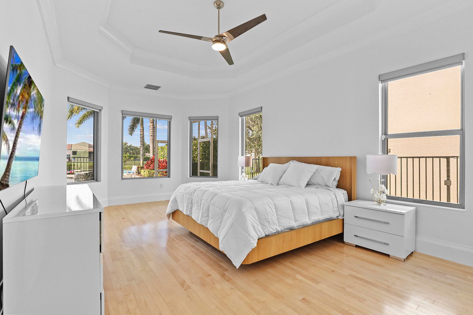 This is a bright and airy primary bedroom featuring a large bed with white linens, light wood flooring, and multiple windows offering natural light. A modern ceiling fan adds to the room's comfort, while a white nightstand and dresser provide storage and style. The overall impression is clean, spacious, and inviting.