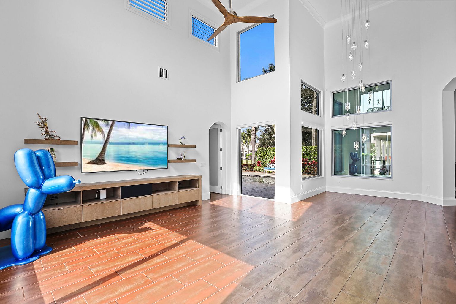 This is a spacious living room featuring high ceilings and an open floor plan. The room is well-lit with natural light streaming through large windows. A modern entertainment center with a large television and floating shelves anchors the space, while the tile flooring adds a touch of elegance. The perspective is from the corner of the room, capturing the full expanse of the living area.