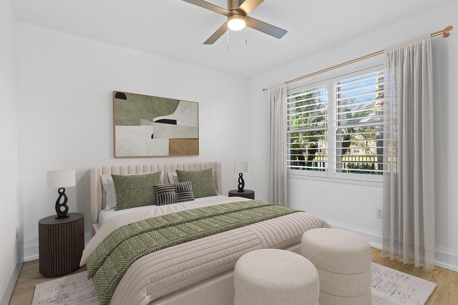 This is a well-staged primary bedroom featuring a neutral color palette with green accents. The room includes a bed with a padded headboard, decorative pillows, and a patterned throw. There is artwork above the bed, side tables with lamps, and two ottomans at the foot of the bed. A window with white shutters and light curtains allows natural light into the room.