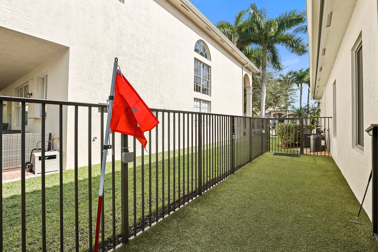This image showcases a well-maintained side yard or garden area, featuring artificial turf and a black metal fence. A red flag marks a putting green, suggesting a recreational space. The backdrop includes a building with stucco siding and palm trees, creating a tropical residential setting.