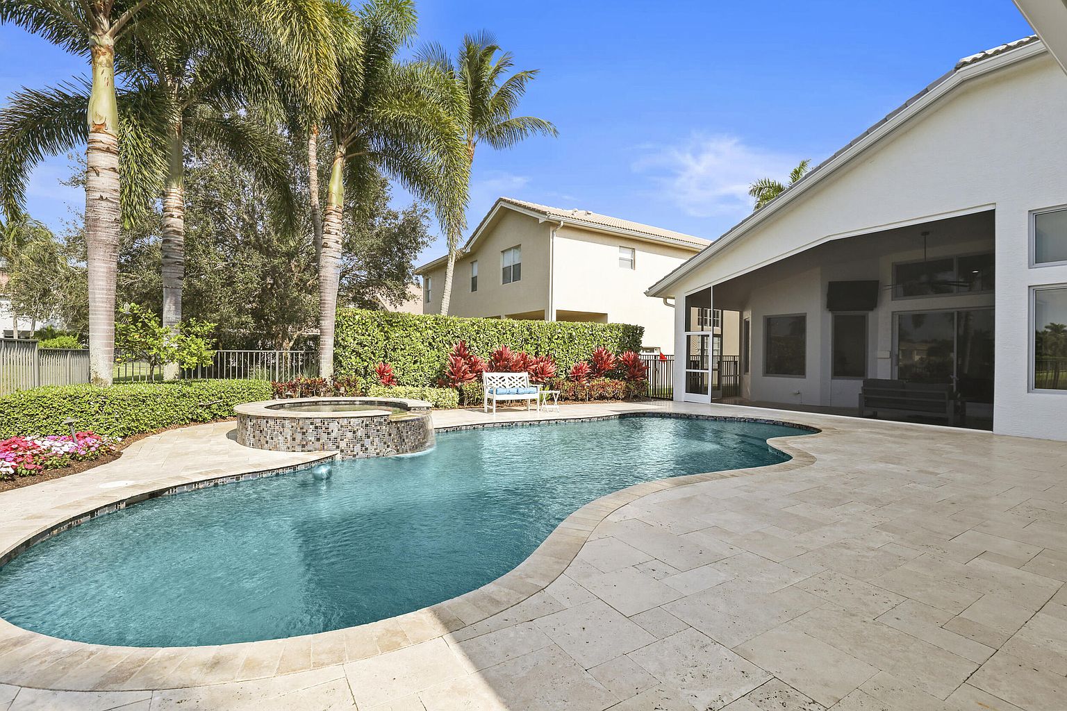 This is a beautiful backyard featuring a freeform pool and spa. The pool is surrounded by a travertine deck and lush landscaping, including palm trees and flowering plants. A covered patio area extends from the house, offering a shaded space for relaxation and entertainment, enhancing the property's appeal for outdoor living.