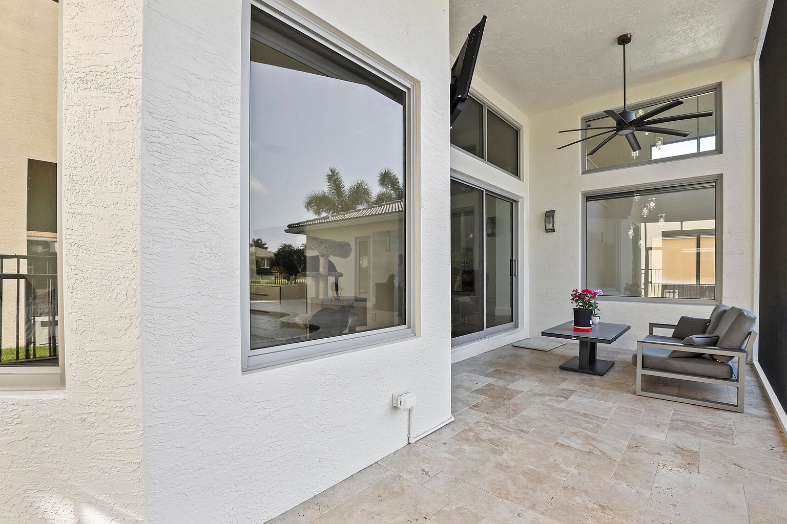 This image showcases a well-appointed outdoor patio area. The space features travertine flooring, a modern outdoor seating arrangement, and a small table with floral decor. Large windows and sliding glass doors provide a seamless transition between indoor and outdoor living, enhancing the appeal of the property.