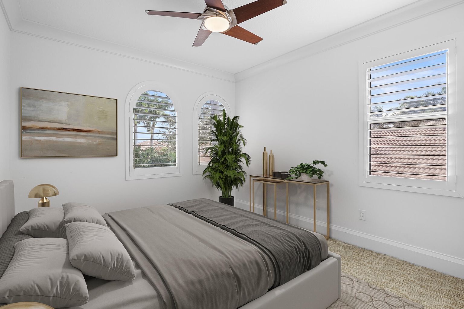 This is a well-lit primary bedroom featuring a neutral color palette. The room includes a large bed with gray bedding, a piece of artwork on the wall, and two arched windows with shutters. A console table with decorative items and a tall potted plant add to the room's decor, while a ceiling fan provides air circulation.