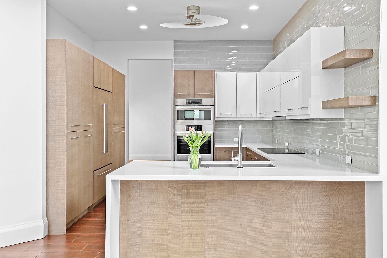This is a well-lit, modern kitchen featuring a large island with a white countertop and light wood paneling. The kitchen includes stainless steel appliances, a combination of white and light wood cabinets, and a light gray tiled backsplash. The flooring appears to be a reddish-brown tile, and a vase of flowers sits on the island, adding a touch of color.