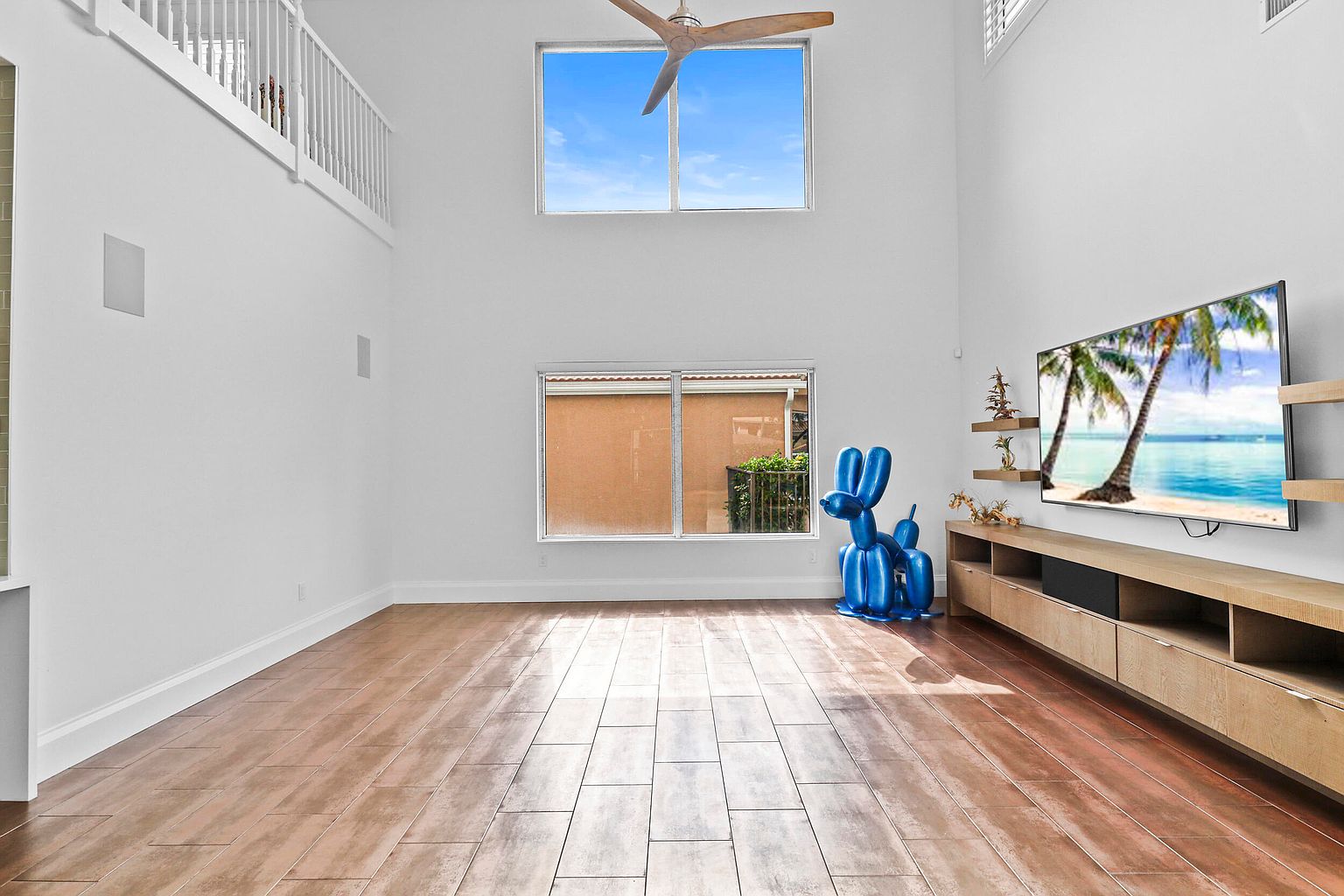 This is a bright and airy living room with high ceilings and large windows that provide ample natural light. The room features wood-look tile flooring, a modern entertainment center with a large television displaying a tropical scene, and a whimsical blue balloon dog sculpture. The overall impression is clean, spacious, and contemporary.