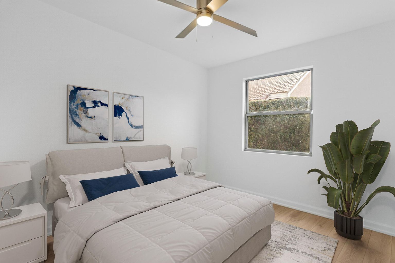 This is a bright and airy primary bedroom featuring a neutral color palette. The room includes a bed with blue accent pillows, two abstract art pieces above the headboard, and a large window providing natural light. A potted plant adds a touch of greenery, enhancing the serene and inviting atmosphere.