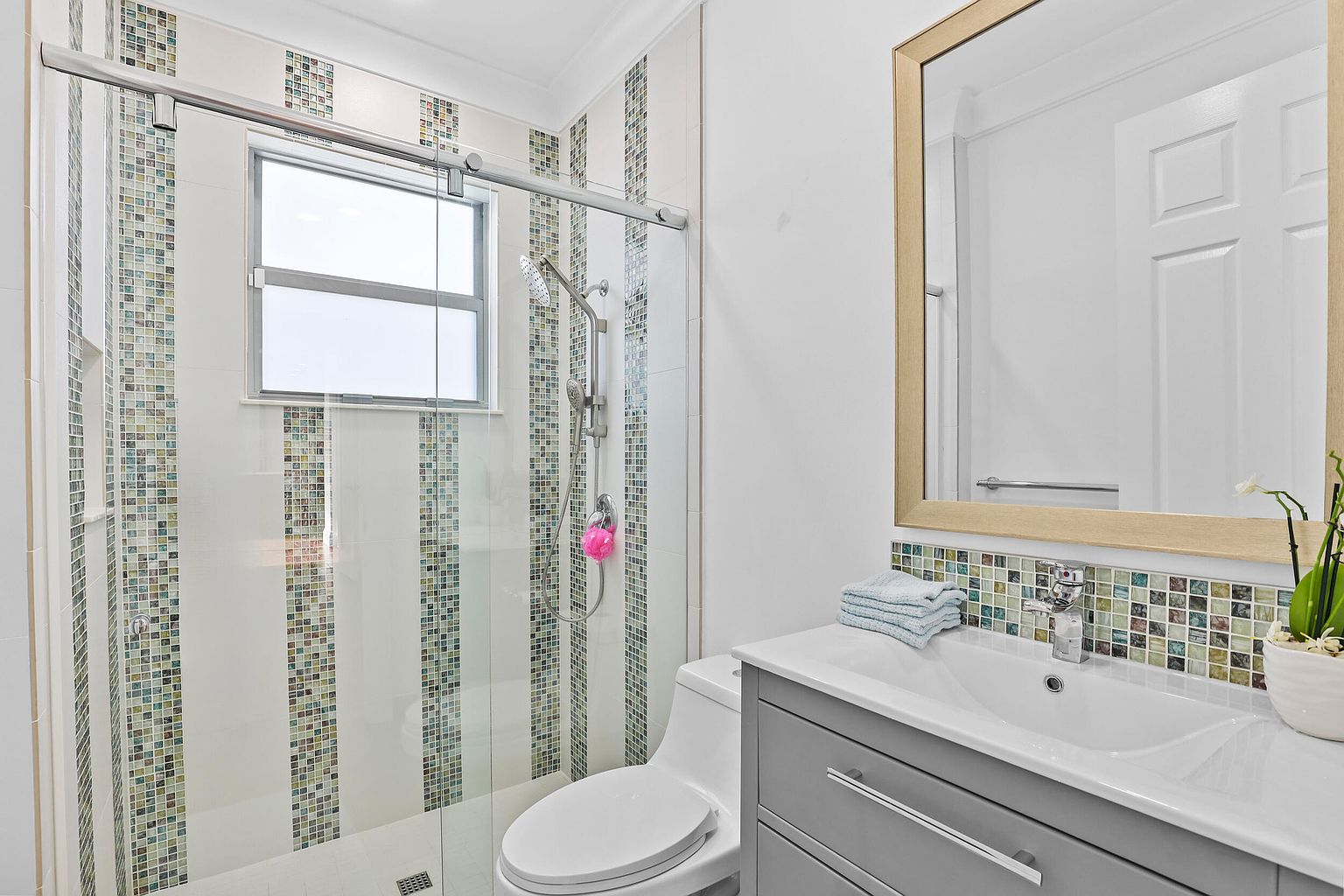 This is a well-lit bathroom featuring a glass-enclosed shower with decorative tile stripes, a toilet, and a vanity with a gray cabinet and a white countertop. A framed mirror hangs above the vanity, reflecting a clean and modern aesthetic. The perspective is from a medium distance, capturing the key elements of the bathroom in a balanced composition.
