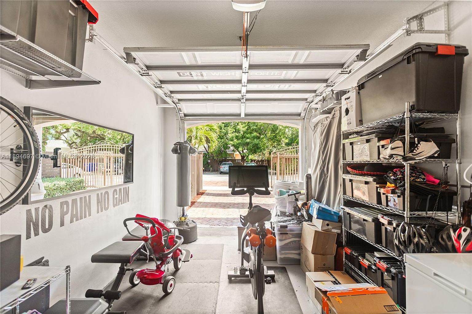 This is an interior shot of a home gym set up inside a garage. The space is equipped with workout equipment such as a stationary bike, punching bag, and weight bench. Storage shelves line one side, and the garage door is partially open, providing natural light and a view to the outside.