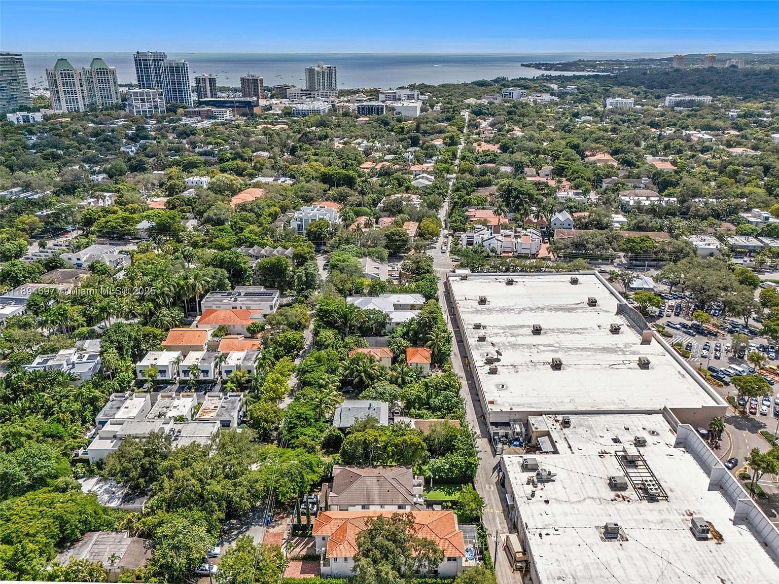 This is an aerial view of a residential area mixed with commercial property featuring lush landscaping and a mix of building styles. The roof of a large commercial building is visible, alongside residential houses and a street with parked cars. In the distance, the cityscape meets the horizon over the ocean.