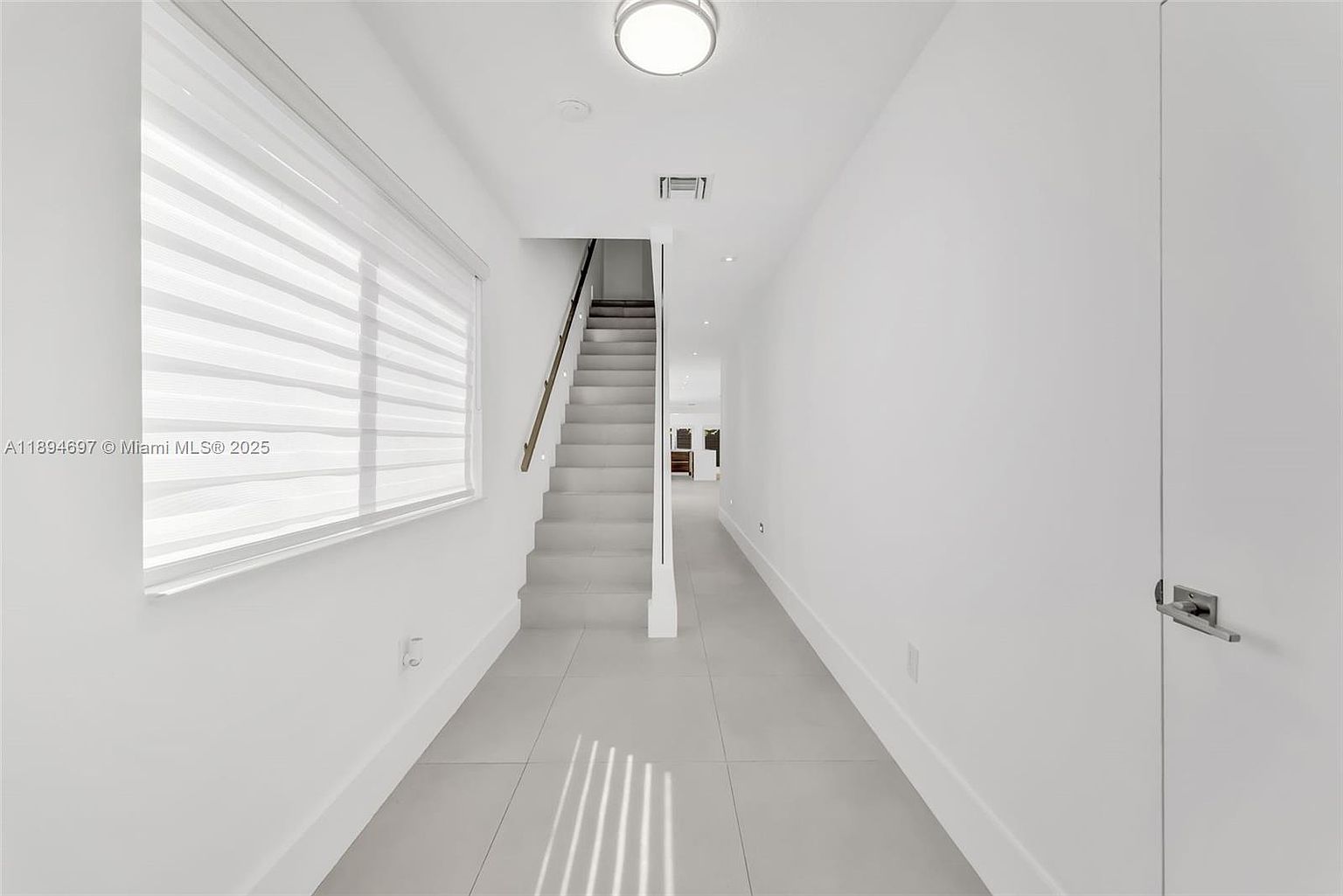 This is an interior shot of a bright, modern hallway with stairs. The walls are painted white, and the floors are covered in light gray tiles. A large window with blinds allows natural light to flood the space, and the staircase leads to an upper level.