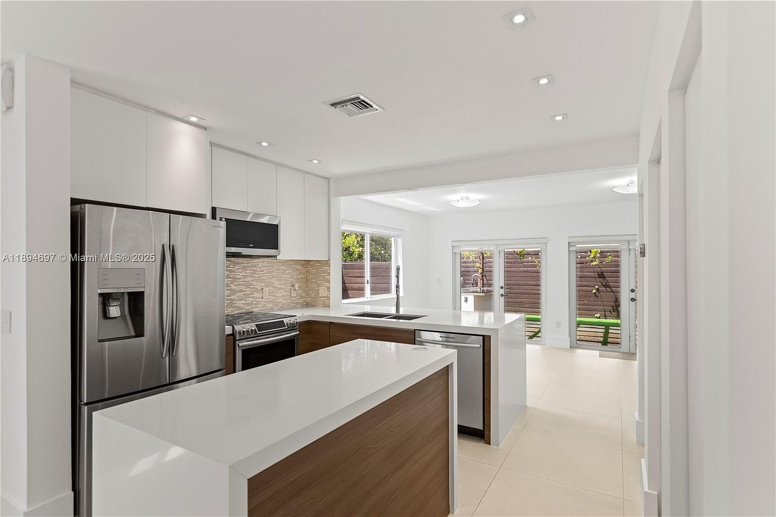 This is a bright and modern kitchen featuring white cabinetry, stainless steel appliances, and a two-tiered island with a white countertop and wood-paneled base. The kitchen has an open layout with a view of the outdoor area through a window and sliding glass doors, creating a seamless indoor-outdoor flow. The flooring is light-colored tile, and recessed lighting illuminates the space.
