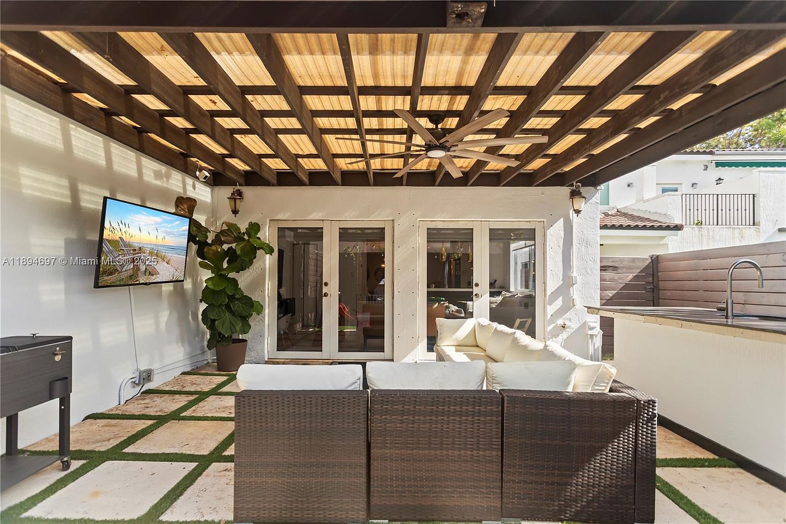 This is a well-appointed outdoor patio area, featuring a woven outdoor sofa with white cushions, set on a stone patio with grass accents. Above, a wooden pergola provides shade, with a ceiling fan for added comfort. A wall-mounted television and an outdoor kitchen area enhance the space for entertainment and relaxation.