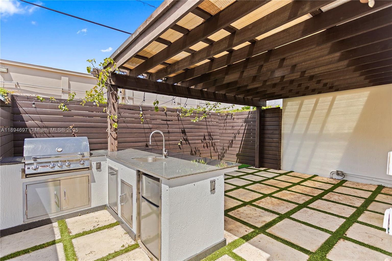 This image showcases an outdoor patio area featuring a built-in stainless steel grill and sink setup, perfect for outdoor cooking and entertaining. The patio is paved with square stones interspersed with grass, creating a visually appealing and functional space. A wooden pergola provides shade, enhancing the ambiance of this outdoor living area.