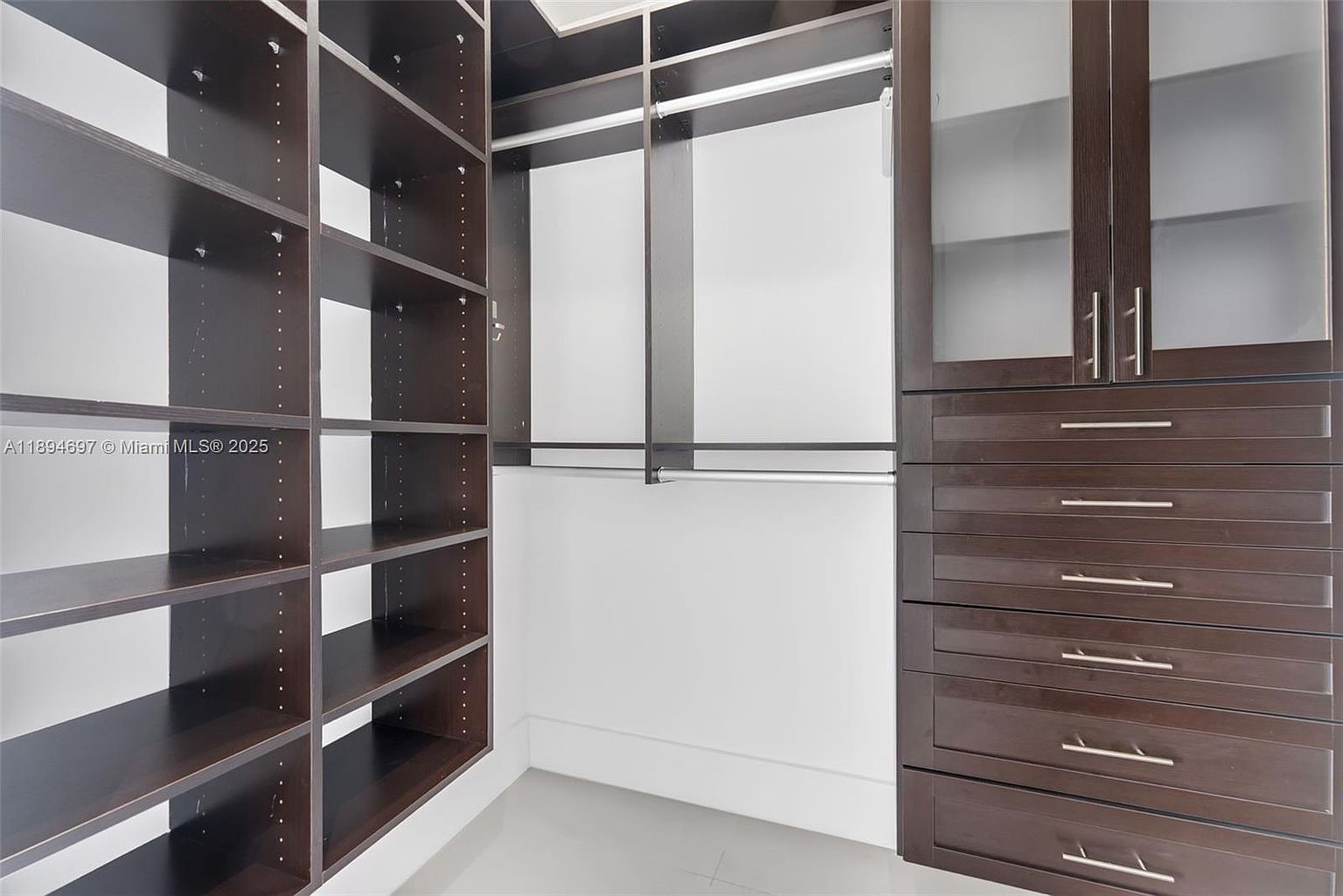 This is a well-organized walk-in closet featuring dark wood shelving and drawers. The closet includes open shelving on the left, hanging rods in the center, and a cabinet with frosted glass doors and drawers on the right. The space is clean and bright, offering ample storage.