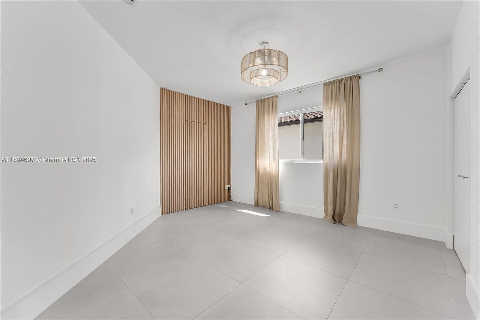 This is a bright and airy bedroom featuring a modern design with clean lines. The room has light gray tile flooring, white walls, and a unique wood slat accent wall. A window with neutral-toned curtains provides natural light, and a stylish light fixture hangs from the ceiling, creating a serene and inviting atmosphere.