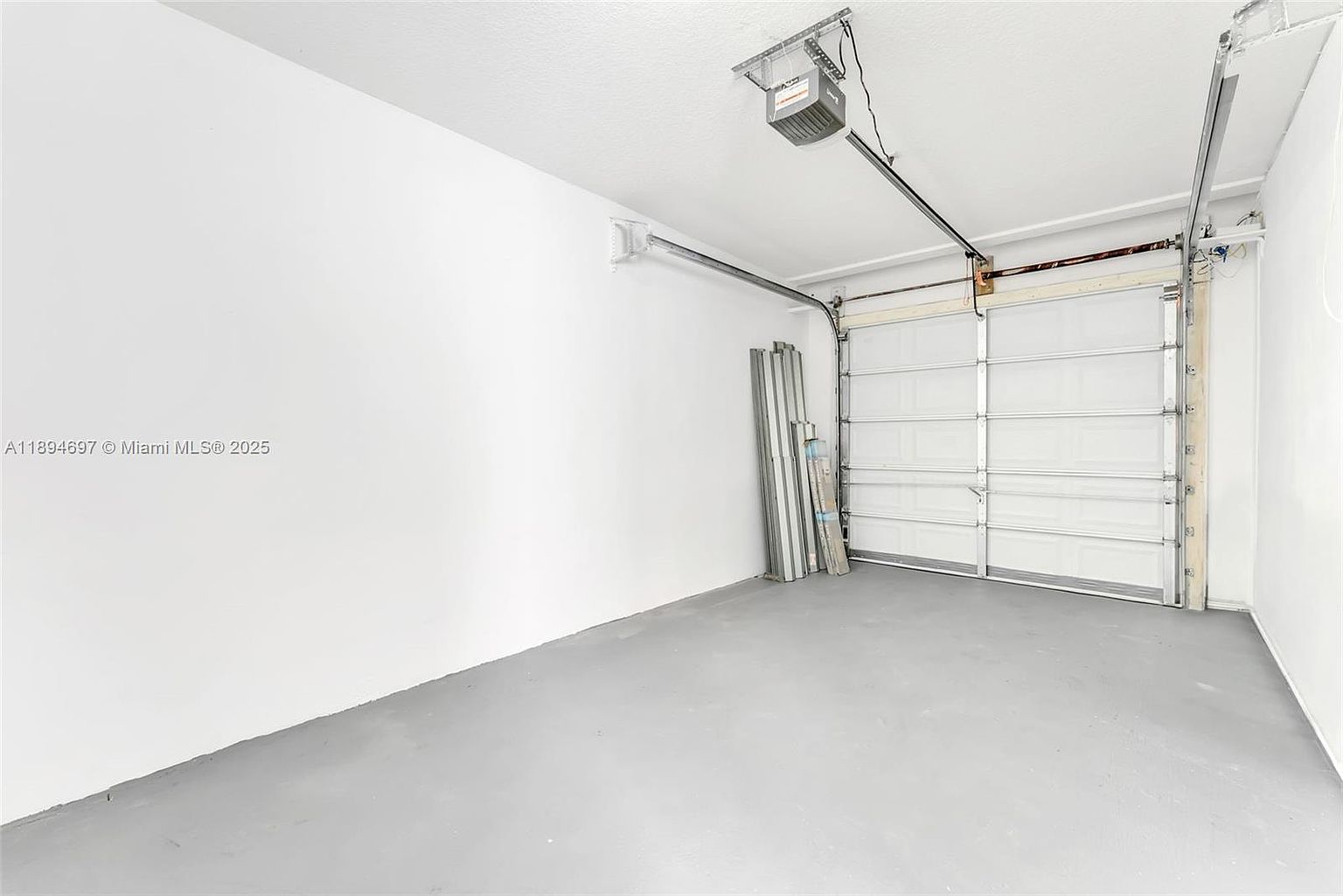 This is an interior shot of a garage. The garage features a white garage door, a gray floor, and white walls. There are some metal objects leaning against the wall near the garage door. The perspective is from the center of the garage, looking towards the door.