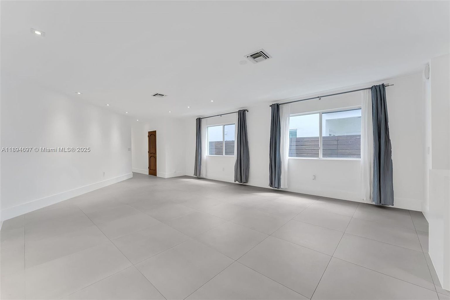 The image showcases a spacious living room with a minimalist design. The room features light gray tile flooring, white walls, and two large windows dressed with gray curtains, allowing ample natural light. A wooden door is visible on the left side of the room, and the overall impression is clean and modern.