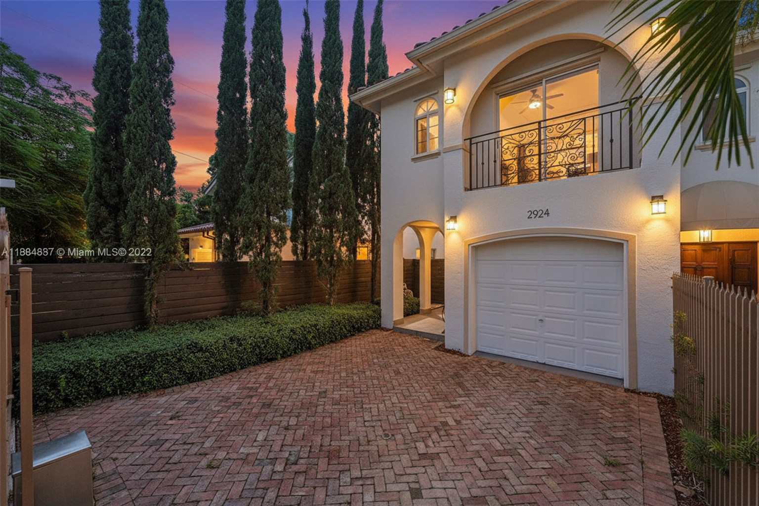 The image showcases the front exterior of a two-story home with a Mediterranean architectural style. Noticeable are the arched entryways, a balcony with decorative iron railings, a two-car garage, and a brick driveway. The facade is painted in a light color, complemented by lush landscaping, including tall slender trees lining the property.
