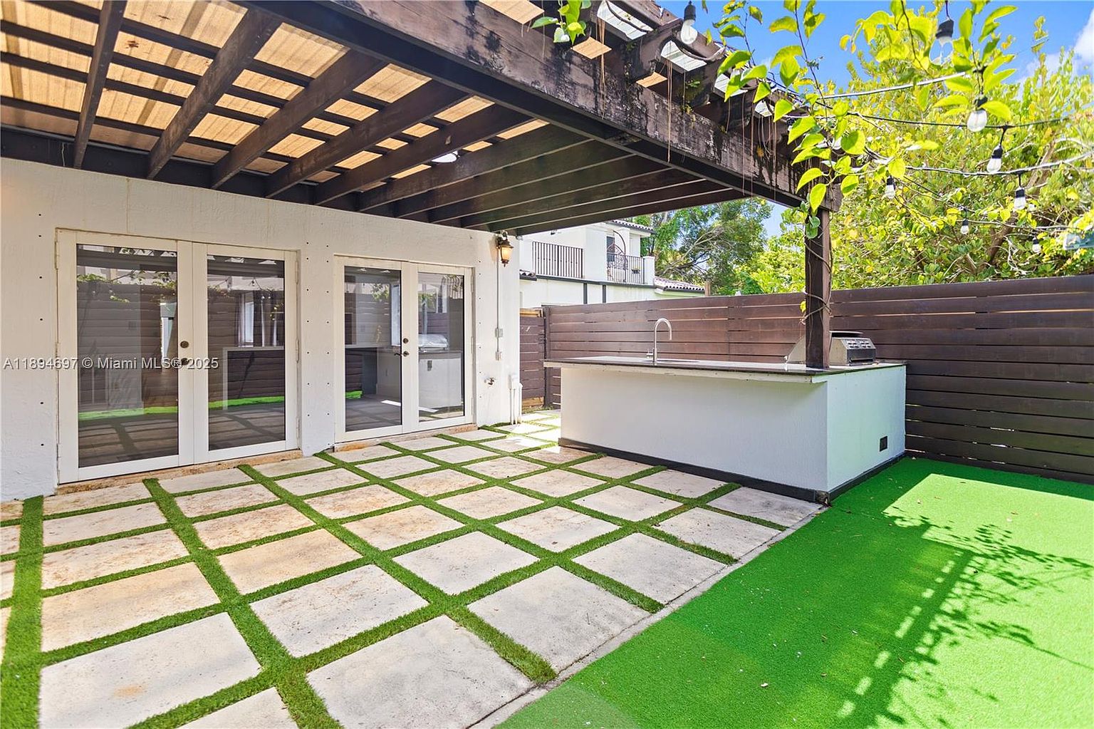 This image showcases a well-designed outdoor patio area, featuring a pergola overhead and a built-in outdoor kitchen with a sink and grill. The patio flooring consists of concrete squares interspersed with artificial turf, creating a modern and inviting space. A wooden fence provides privacy, and string lights add a touch of ambiance.