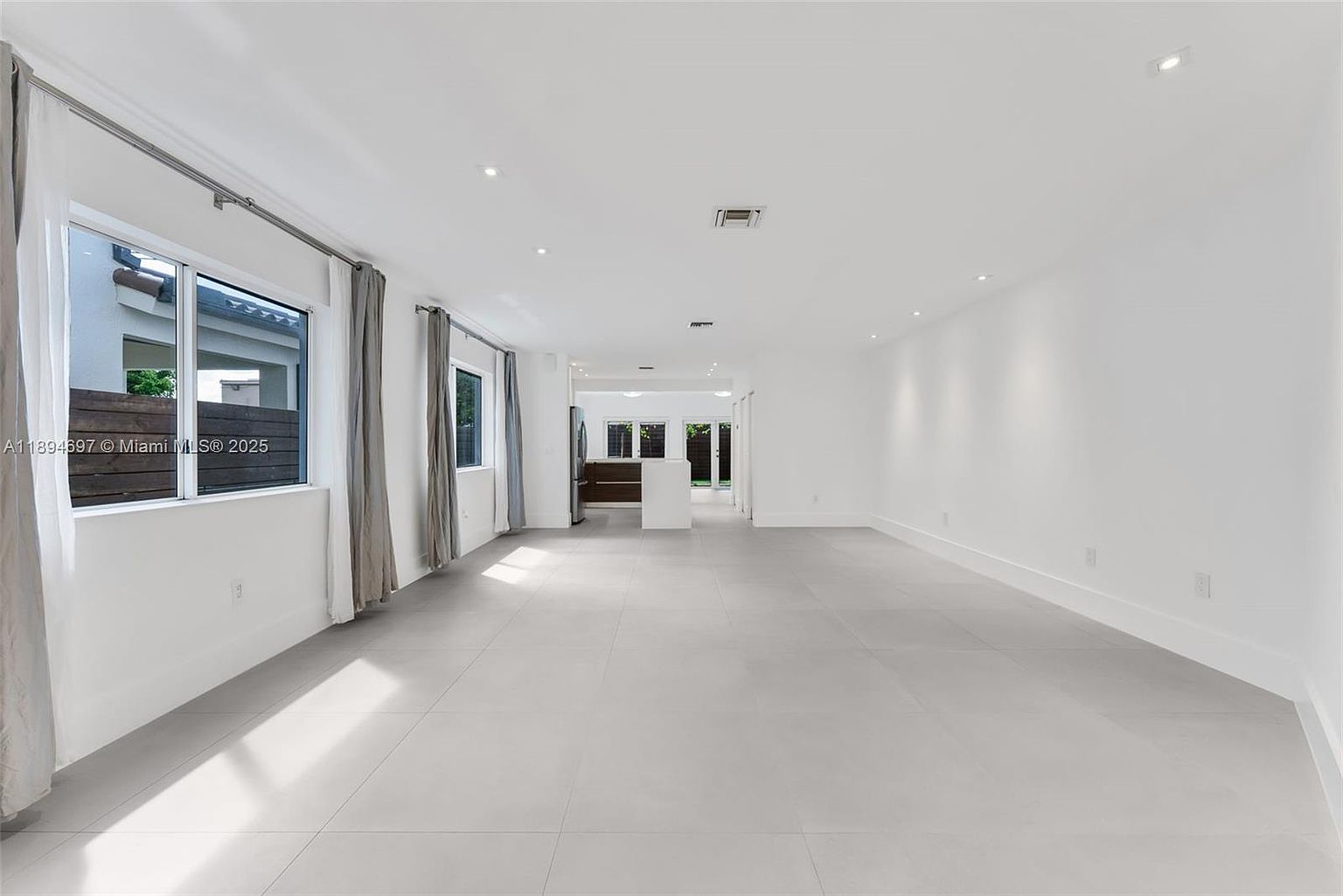 This is a bright and spacious living room featuring neutral-toned tile flooring and white walls, creating a modern and minimalist aesthetic. Large windows with light-colored curtains provide ample natural light, enhancing the open feel of the space. The room appears to be part of an open-concept design, leading into other areas of the home.