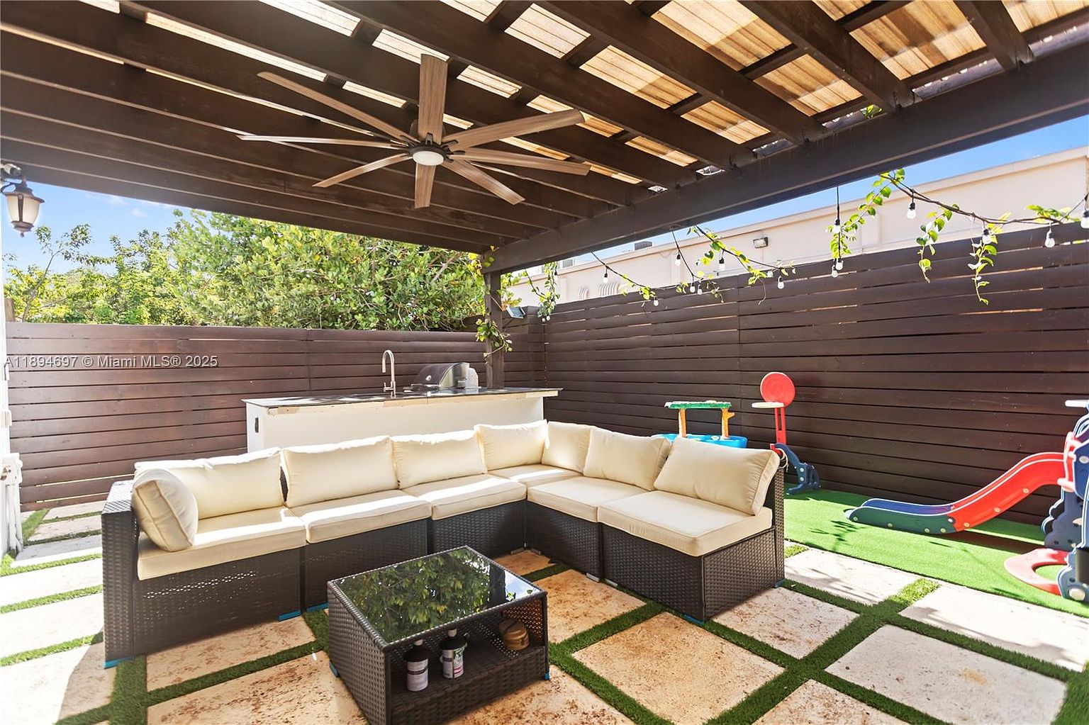 This is a cozy outdoor patio area featuring a large sectional sofa with light cushions, a dark wicker coffee table, and a built-in outdoor kitchen area with a sink and grill. The patio is covered by a pergola with a ceiling fan, and the flooring is a combination of stone tiles and artificial turf. A wooden fence provides privacy, and string lights add a warm ambiance.
