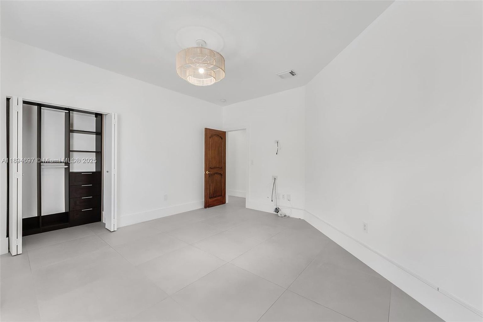 The image showcases a bright, empty bedroom with a minimalist aesthetic. The room features light gray tile flooring, white walls, a modern light fixture, and a dark wood closet with sliding doors. A wooden door leads to another room, and the overall impression is clean and spacious, ready for personalization.