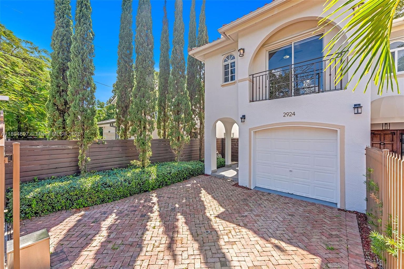 This is a front exterior view of a two-story Mediterranean-style home. The house features a white stucco facade, a white garage door, and a small balcony with decorative iron railings. A brick driveway leads up to the garage, and tall, slender trees line the side of the property, adding to the curb appeal.