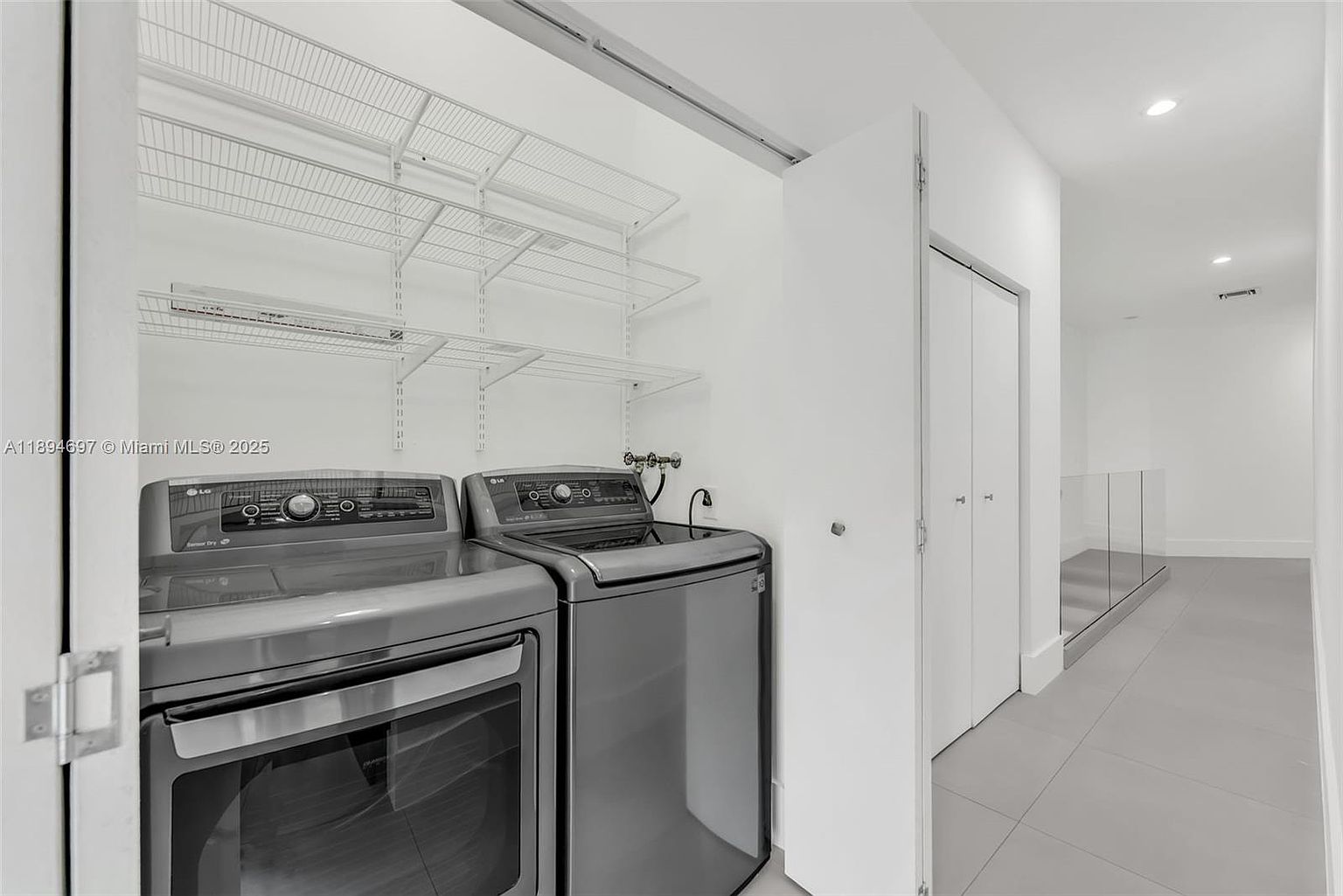This is an interior shot of a laundry room featuring a gray front-load washer and dryer set. Above the appliances are wire shelving units for storage. The room is painted in a neutral color, and there is a doorway leading to a hallway with a glass railing.