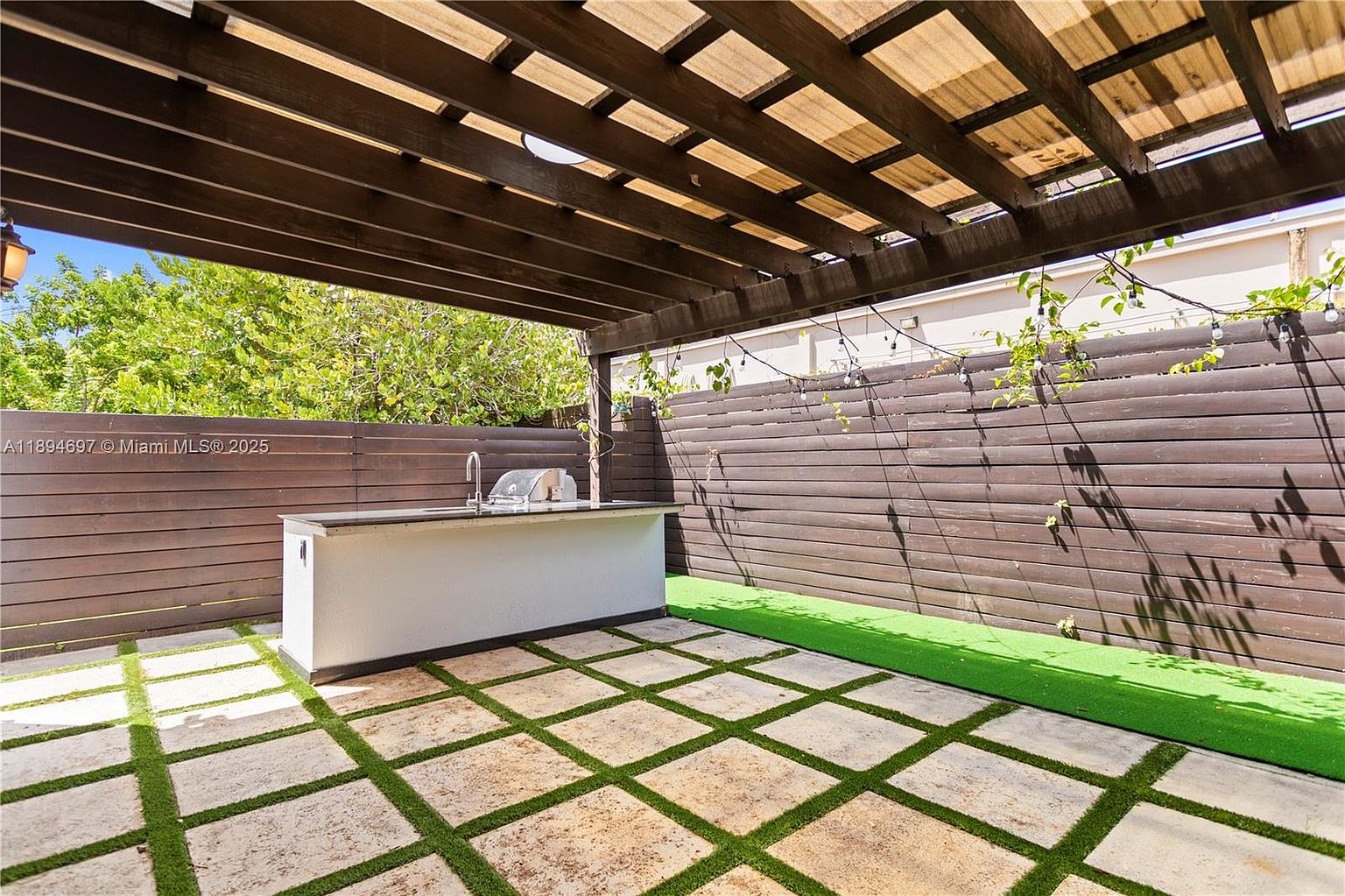 This is a well-designed outdoor patio area featuring a built-in grill station with a sink, perfect for entertaining. The flooring consists of square pavers with artificial turf in between, adding a touch of greenery. A wooden pergola provides shade, and string lights add a cozy ambiance to the space.