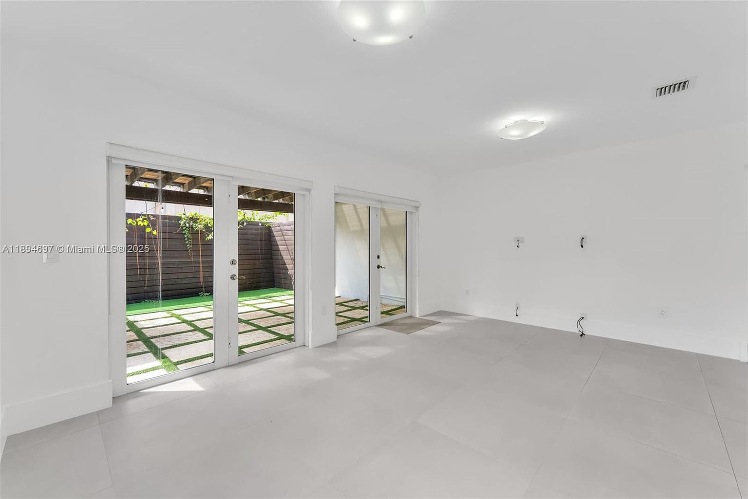 The image showcases a bright and airy living room with white walls and light gray tile flooring. Large sliding glass doors provide access to an outdoor patio area with artificial turf, enhancing the indoor-outdoor flow. The room is minimalist in style, emphasizing clean lines and natural light.