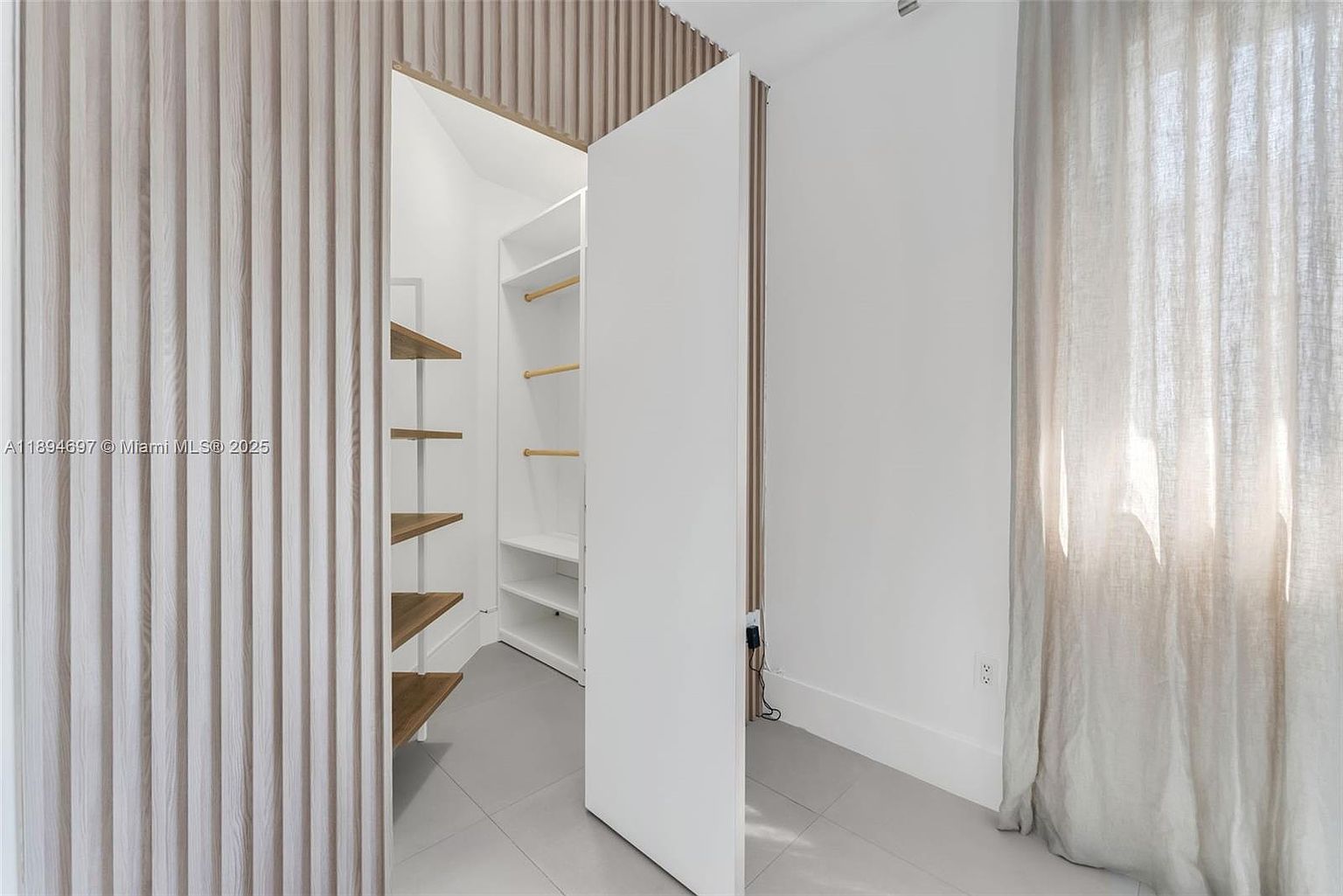 This image showcases a modern walk-in closet with a unique design. The closet features a combination of open shelving with wooden accents and closed storage behind a large white door. The interior is well-lit, and the overall aesthetic is clean and minimalist, creating a sense of organized luxury.