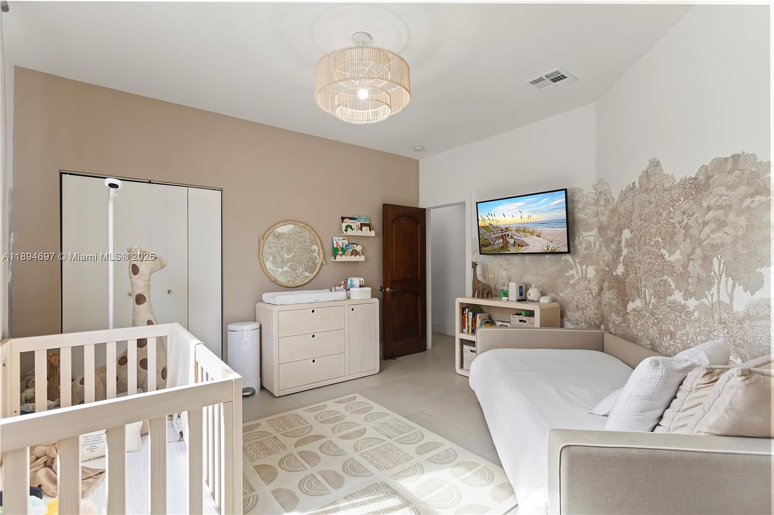 This is a cozy nursery featuring a crib, changing table with drawers, and a daybed. The room is decorated in neutral tones with a light wood chandelier and a patterned rug. A TV is mounted on the wall above a small bookshelf, and a tree-themed wallpaper adds a whimsical touch.