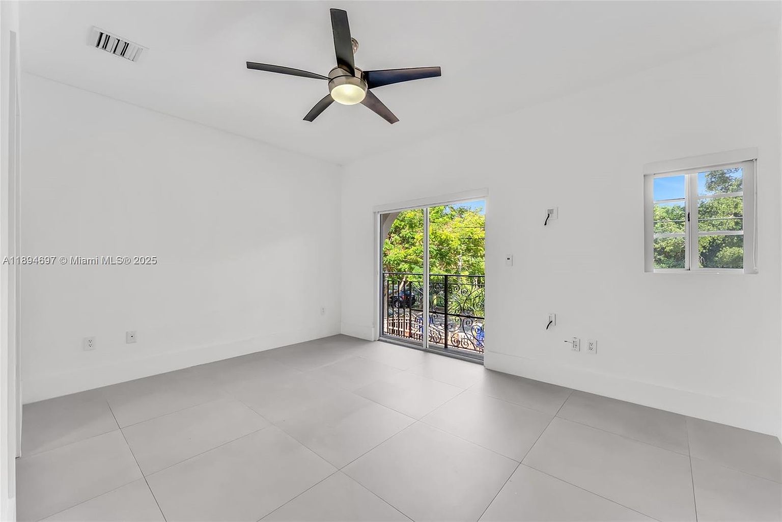 The image showcases a bright, minimalist bedroom with white walls and light gray tile flooring. A ceiling fan is centrally located, and natural light streams in through a sliding glass door leading to a balcony and a window on the right. The room is spacious and uncluttered, presenting a clean and modern aesthetic.