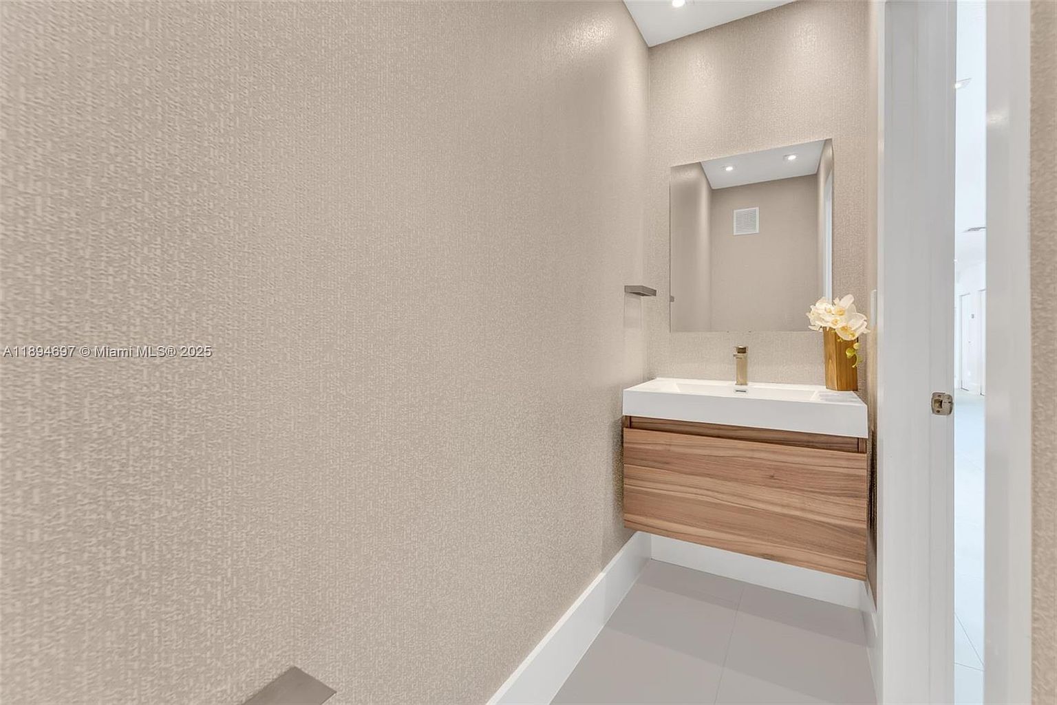 This is a modern guest bathroom featuring a floating vanity with a light wood finish and a white countertop. A rectangular mirror hangs above the sink, and the walls are covered in a textured beige wallpaper. The floor is tiled in a light gray, and a doorway to the right offers a glimpse into another room, creating a clean and contemporary aesthetic.