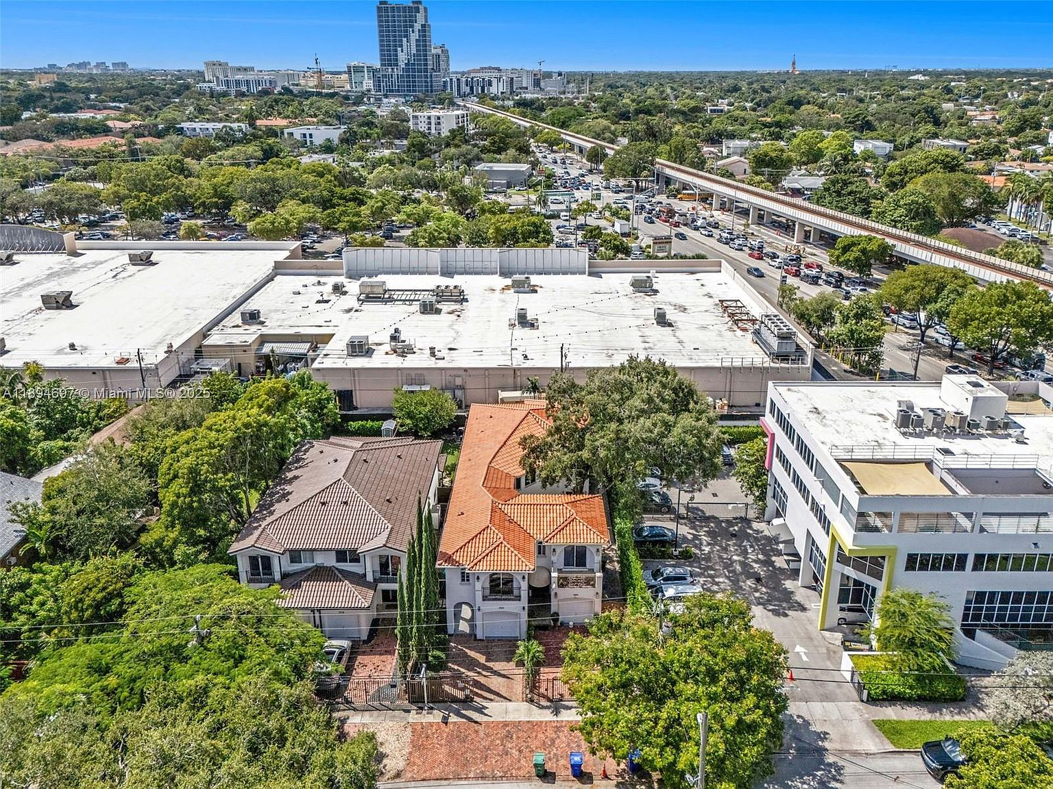 This aerial shot captures a residential area, showcasing homes with reddish-brown tiled roofs situated near a large retail building with a white roof. The perspective highlights proximity to commercial areas and transportation infrastructure, including roads and potentially a train line. Lush greenery surrounds the buildings, creating a mix of urban and suburban elements in the scene.