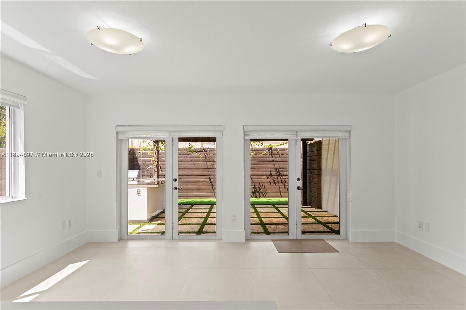 This is an interior shot of a living room featuring white walls and tile flooring. Two sets of glass doors lead to an outdoor patio area with a wooden fence and artificial turf. The room is illuminated by two ceiling lights, creating a bright and airy atmosphere.