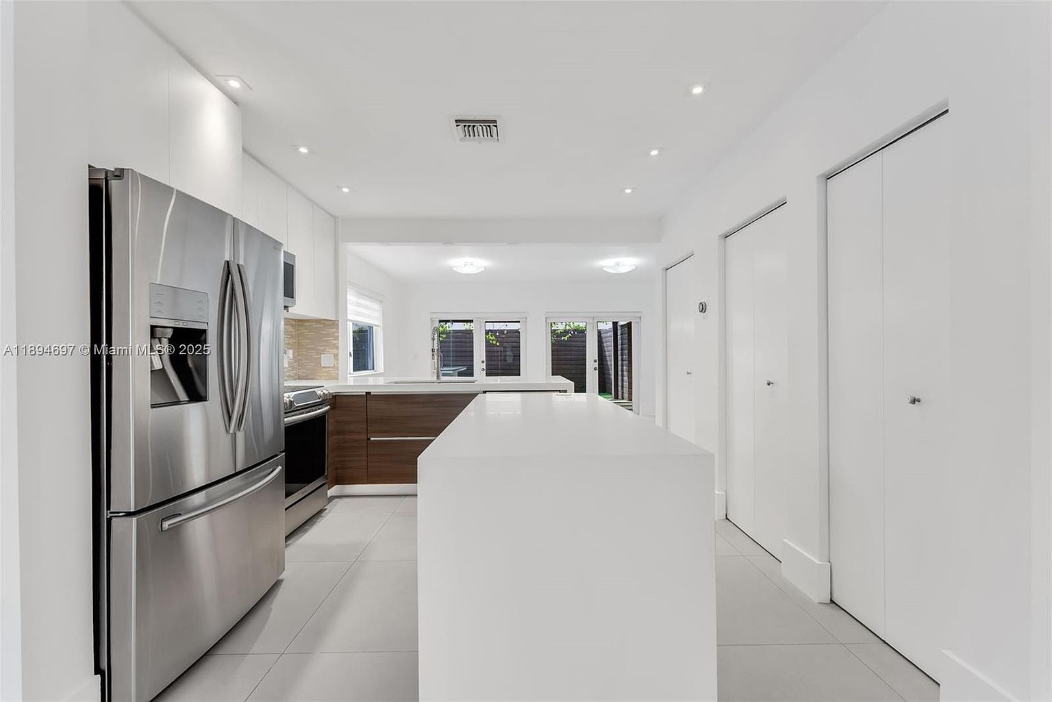 This is a bright and modern kitchen featuring stainless steel appliances, white cabinetry, and a large white island. The kitchen has an open layout with a view into another room through a wide opening. The flooring is light gray tile, and the overall impression is clean and spacious.