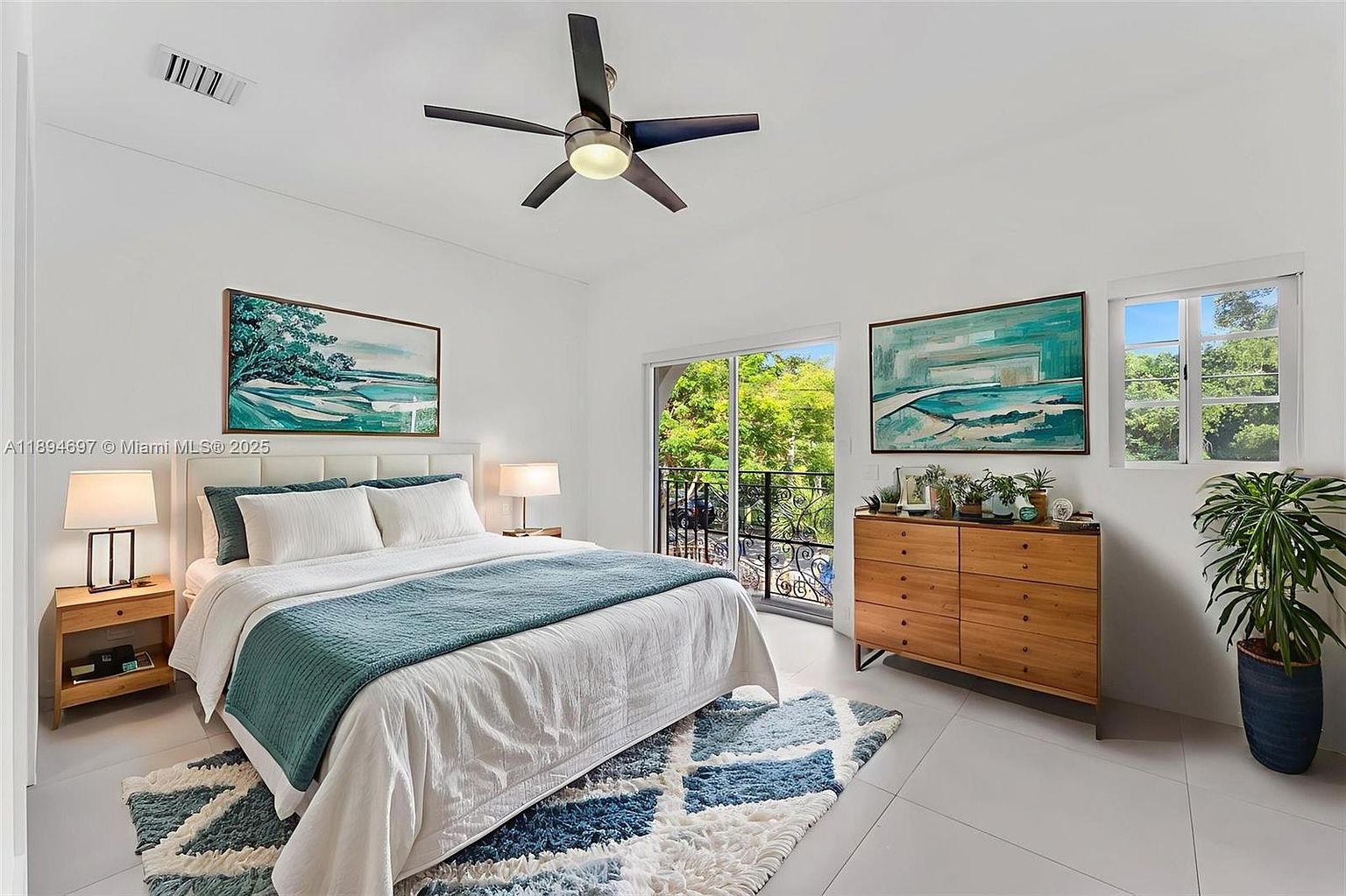 This is a bright and airy primary bedroom featuring a large bed with white linens and a teal throw, complemented by matching artwork. The room has a modern aesthetic with a ceiling fan, light wood dresser, and a balcony door that lets in natural light. A blue and white patterned rug adds a touch of color and texture to the space.
