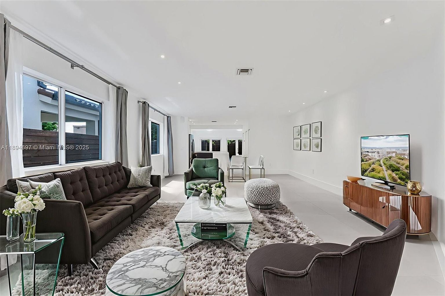 This is a bright and spacious living room featuring a brown sofa, a green armchair, and a marble-topped coffee table on a shaggy rug. The room is well-lit with natural light from a large window and recessed lighting, complemented by a modern TV and console. The overall impression is clean, contemporary, and inviting.